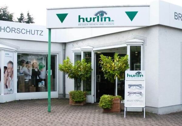 Eingangsbereich eines Optiker- und Hörakustik-Fachgeschäfts mit Werbeschildern und Pflanzen.