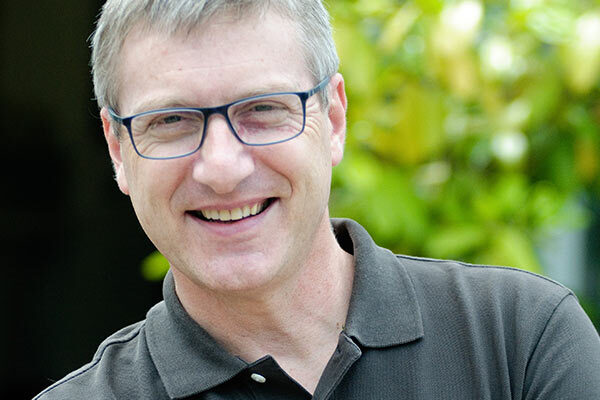 Mann mit Brille und grauen Haaren lächelt freundlich im Garten bei natürlichem Licht.